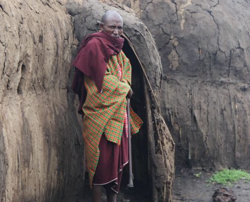 Während Ihrer 7 Tage Safari in Tansania werden Sie die Gelegenheit haben, die Menschen und die Kultur des Landes kennenzulernen, so wie diesen älteren Maasai, der vor einer traditionellen Boma aus Lehm steht.